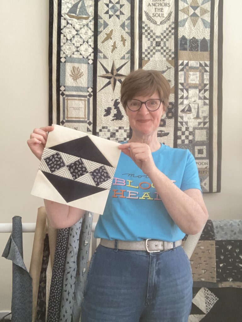 Janet Clare holding her 'Shiny Bauble' block with her 'Spoondrift' quilt in the background.
